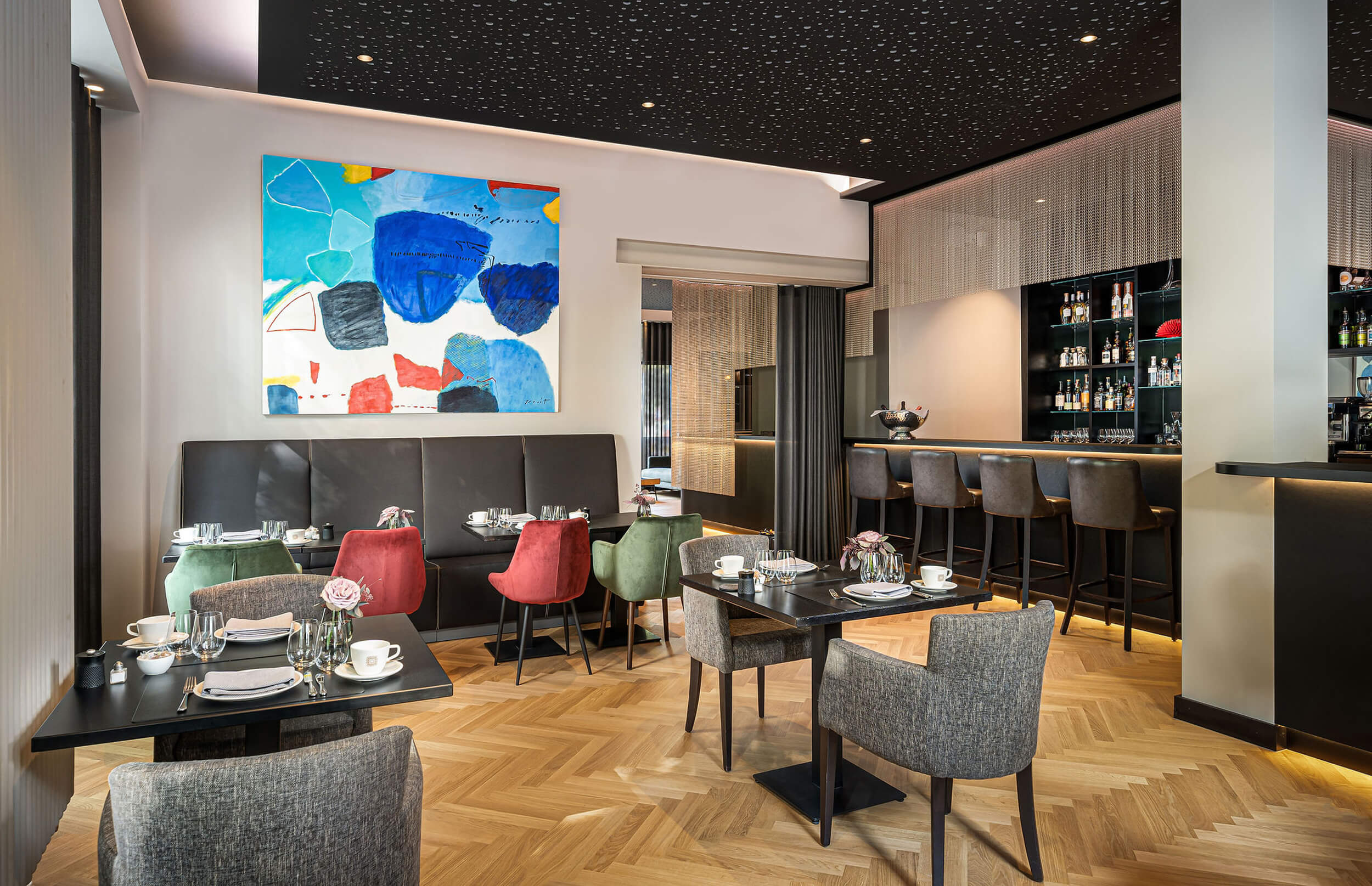 Gedeckte Tische im Fruehstueckssalon. Es zeigt den Fruehstueckssalon mit zum Fruehstueck gedeckten Tischen auf der linken Seite und einen Ausschnitt der Bar auf der rechten Seite. In der Mitte ist der Durchgang zu sehen. Die Datei ist ein Foto im JPEG-Format.