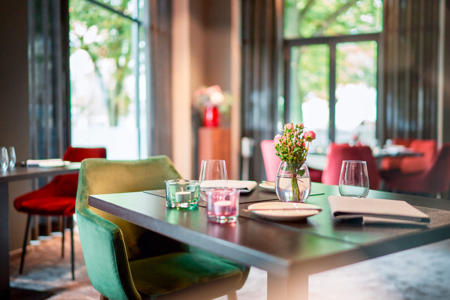 Gourmet-Restaurant ESPLANADE – Tisch. Es zeigt im Vordergrund einen gedeckten Tisch im ESPLANADE Gourmet-Restaurant. Der Hintergrund ist unscharf und weitere Tische sowie grosse Fenster mit einem Ausblick ins Gruene sind darin zu sehen. Die Datei ist ein Foto im JPEG-Format.