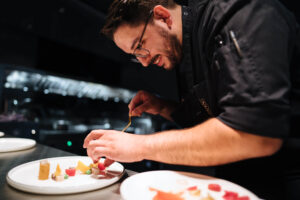 Chefkoch Silio Del Fabro beim Anrichten in der Kueche des Restaurants. Es zeigt rechts Chefkoch Silio Del Fabro der lächelnd auf den Teller auf der Theke links im Bild schaut und das Gericht darauf mit beiden Haenden anrichtet. Die Datei ist ein Foto im JPEG-Format.